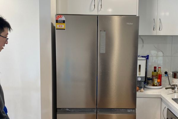 A technician from Quality Appliance Repairs (QAR) inspects a modern Haier French door refrigerator in a well-lit kitchen. The fridge is built into cabinetry and surrounded by various kitchen appliances and utensils, indicating a home service visit.